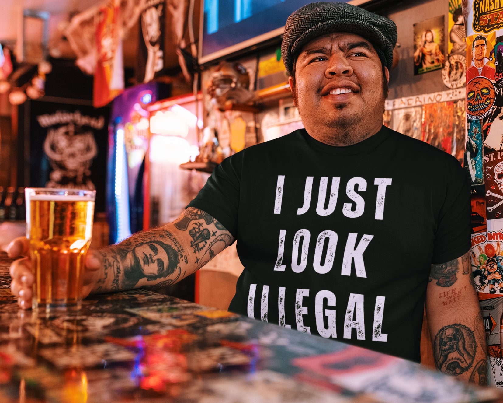 Mexican man standing behind a bar and holding a glass of beer.  The man is wearing a funny black t-shirt for Latinos and Mexican Americans.  The faded, vintage-style lettering reads "I just Look Illegal"