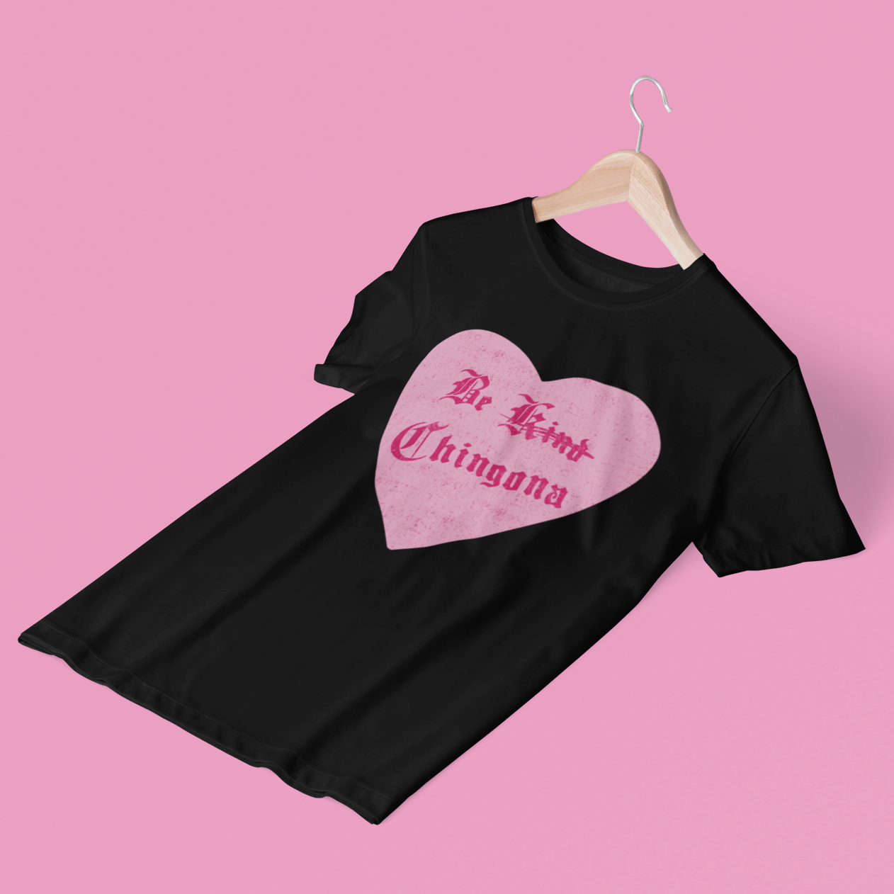 Funny and bold black t-shirt for Latina women with the phrase 'be kind chingona', with the word 'kind' crossed out, in a pink heart graphic and old-English style lettering
