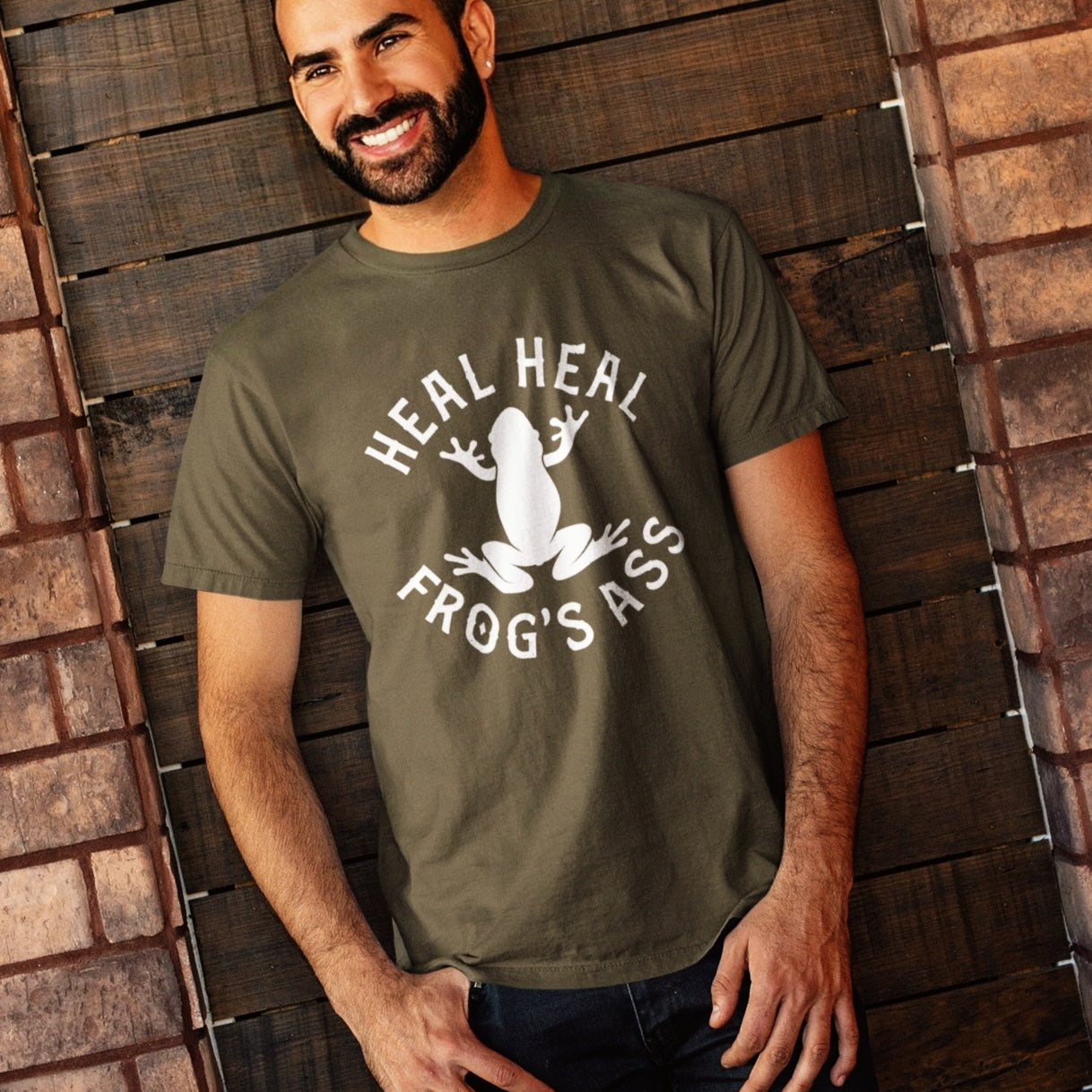 hispanic man wearing a funny navy green t-shirt for Latino culture. the graphic depicts a frog and the words "heal heal frog's ass" in honor of sana sana colita de Rana
