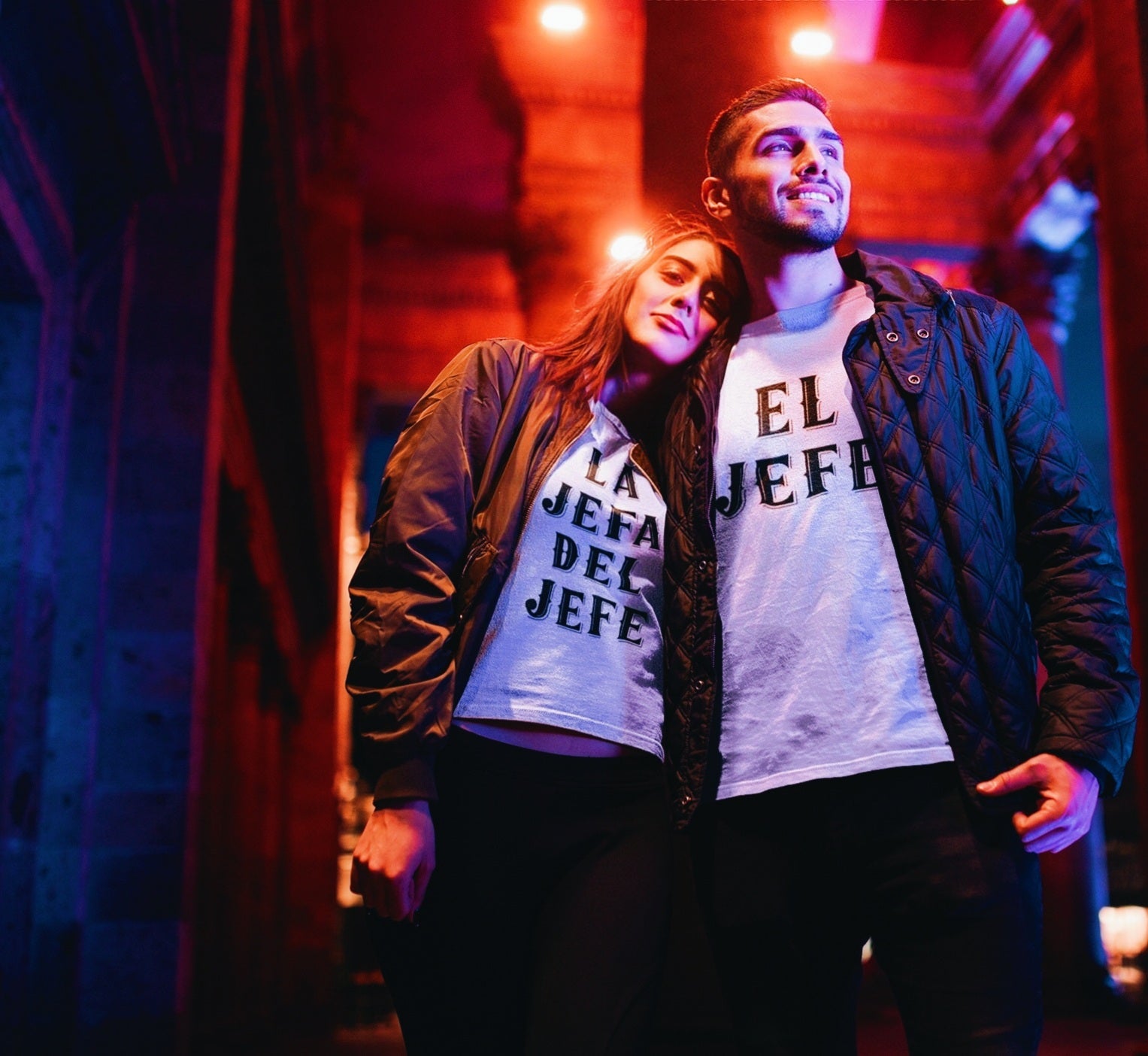 hispanic man and woman wearing funny white t-shirts. the mans's shirt says 'el jefe' and the woman's shirt says 'la jefa del jefe'