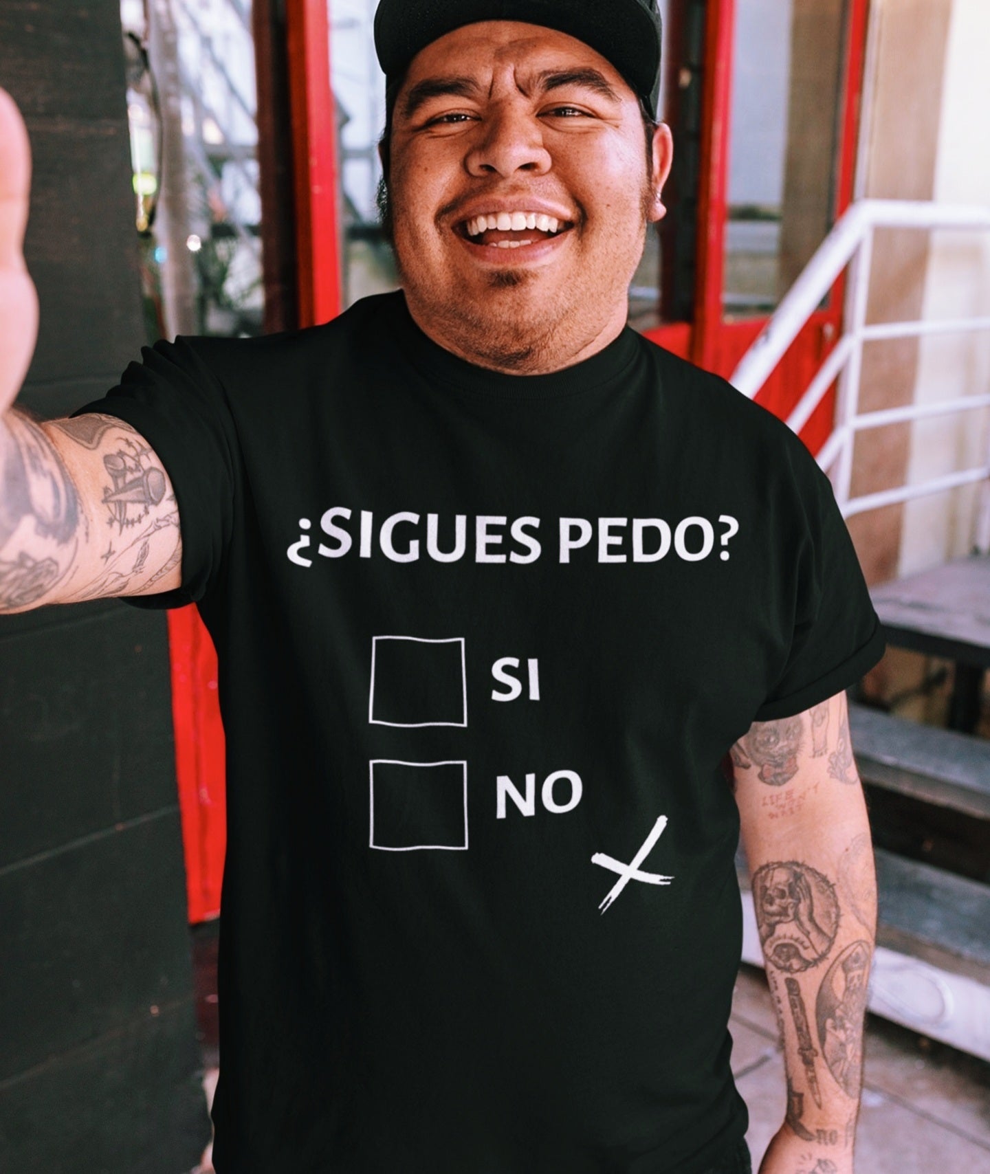Latino man wearing a black T-shirt with text "sigues pedo?" and checkboxes "si" and "no", "no" box attempted to be marked with an "x" but off center, symbolizing a drunken state