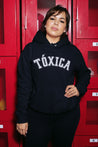 Latina woman wearing a black hoodie sweatshirt with the word 'Toxica' written in white vintage arch-style lettering