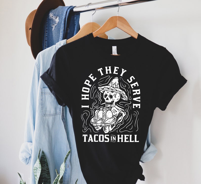Fun and edgy black t-shirt for latina women with a graffiti skeleton illustration wearing a sombrero and smoking a cigarette while serving tacos with the words 'I hope they serve tacos in hell.'