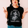 latina wearing a Black t-shirt that showcases a humorous and alternative skeleton graphic serving tacos and smoking a cigarette with the caption 'I hope they serve tacos in hell.'